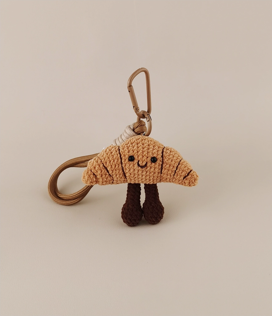 This is a crocheted keychain. It's designed to look like a cute croissant with a smiling face. The croissant has a light brown, textured appearance, black eyes, and a small curved smile. It also features two dark brown, cylindrical legs. Attached to it is a brown cord and a metal carabiner, making it suitable for hanging keys or decorating bags.