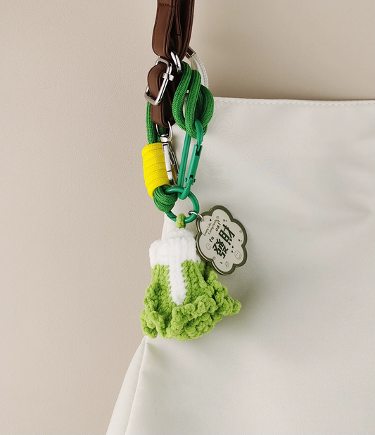 This is a crocheted keychain shaped like a Chinese cabbage. The cabbage has a white stem and green leaves, crafted with fine crochet work. It’s attached to a green carabiner and a green lanyard, with a small tag on the lanyard. The overall design is cute and vivid, making it a lovely accessory for keys or bags.