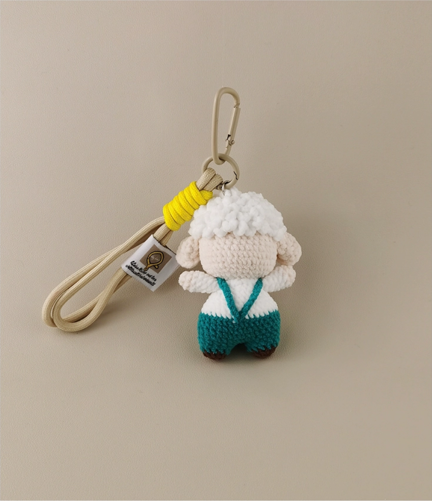 This is a crocheted keychain featuring a cute sheep character. The sheep has a fluffy white "wool" head, a light - colored face and body, and is dressed in teal overalls. It's attached to a beige lanyard with a yellow accent and a metal carabiner. There's also a small tag on the lanyard. The overall design is adorable and handmade - looking.