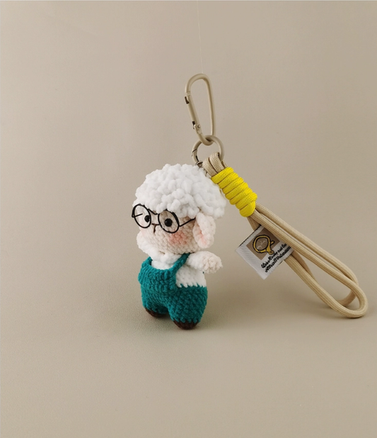 This is a crocheted keychain featuring a cute sheep character. The sheep has a fluffy white "wool" head, a light - colored face and body, and is dressed in teal overalls. It's attached to a beige lanyard with a yellow accent and a metal carabiner. There's also a small tag on the lanyard. The overall design is adorable and handmade - looking.