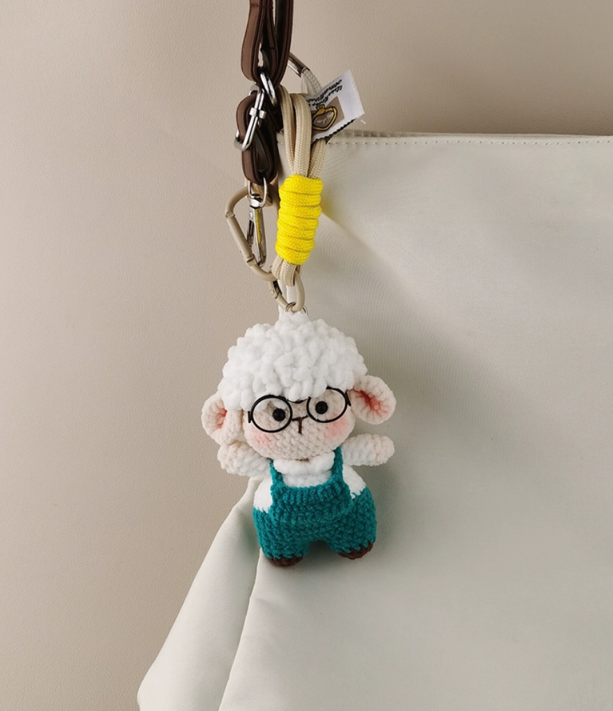 This is a crocheted keychain featuring a cute sheep character. The sheep has a fluffy white "wool" head, a light - colored face and body, and is dressed in teal overalls. It's attached to a beige lanyard with a yellow accent and a metal carabiner. There's also a small tag on the lanyard. The overall design is adorable and handmade - looking.