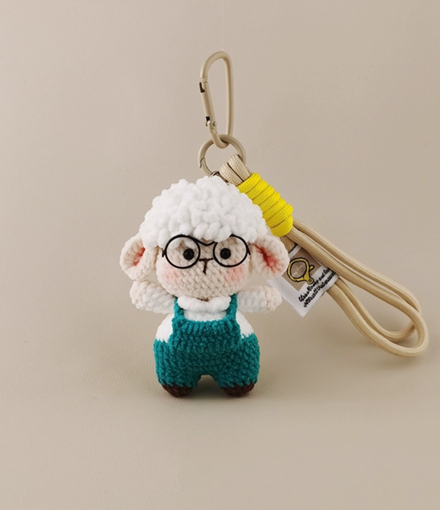 This is a crocheted keychain featuring a cute sheep character. The sheep has a fluffy white "wool" head, a light - colored face and body, and is dressed in teal overalls. It's attached to a beige lanyard with a yellow accent and a metal carabiner. There's also a small tag on the lanyard. The overall design is adorable and handmade - looking.