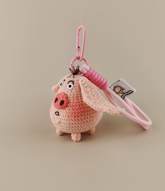This is a crocheted keychain in the shape of a cute pink pig. The pig has large, expressive eyes, a round pink nose, and a small tuft of hair on its head. It's attached to a pink carabiner and a pink lanyard, with a small tag on the lanyard. The overall design is adorable and handmade - looking.
