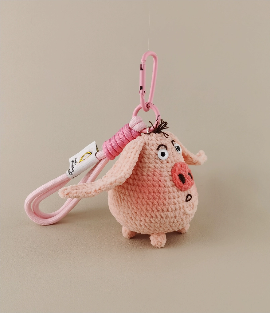 This is a crocheted keychain in the shape of a cute pink pig. The pig has large, expressive eyes, a round pink nose, and a small tuft of hair on its head. It's attached to a pink carabiner and a pink lanyard, with a small tag on the lanyard. The overall design is adorable and handmade - looking.