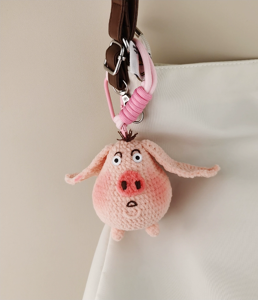 This is a crocheted keychain in the shape of a cute pink pig. The pig has large, expressive eyes, a round pink nose, and a small tuft of hair on its head. It's attached to a pink carabiner and a pink lanyard, with a small tag on the lanyard. The overall design is adorable and handmade - looking.