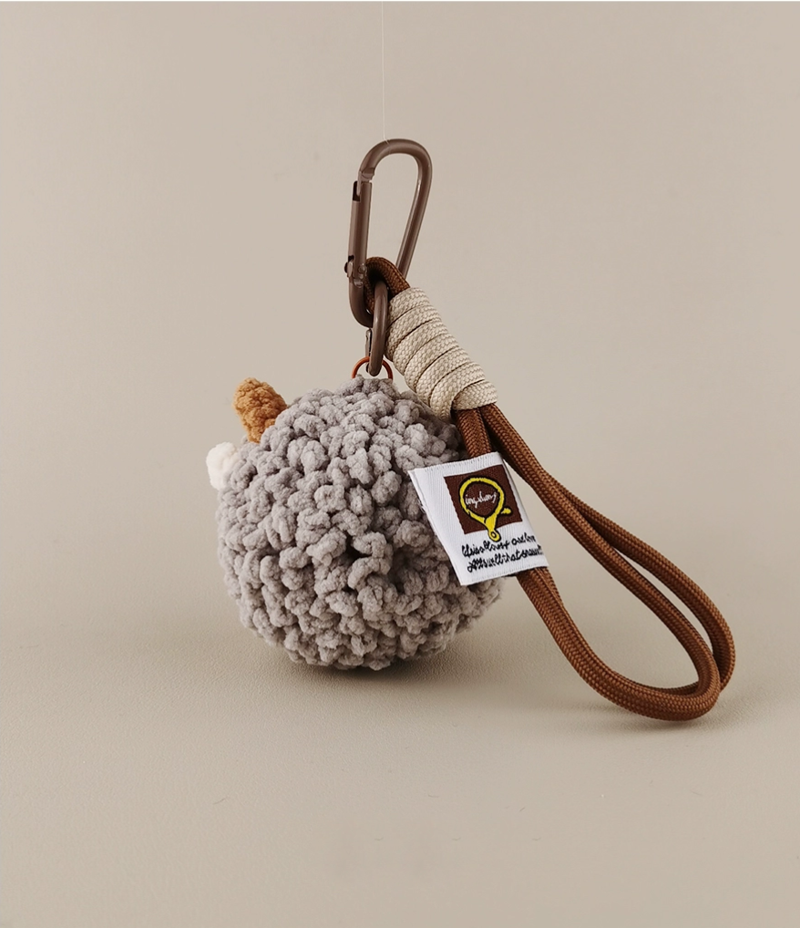 A cute crochet sheep keychain with a tag and brown leather string, on a light gray background, product photography.
