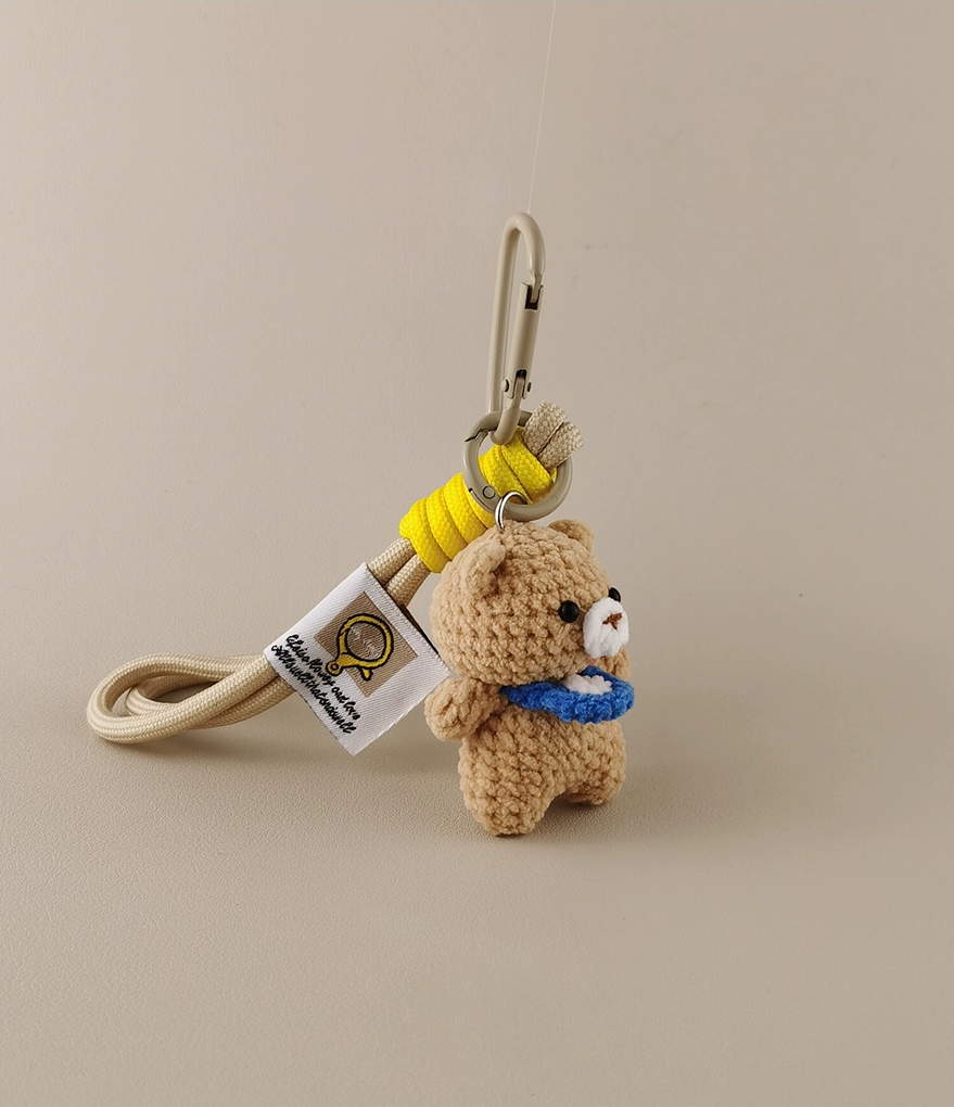 A keychain with an amigurumi brown bear holding a blue paper tag, a yellow lanyard, and a white badge. The background is light gray.