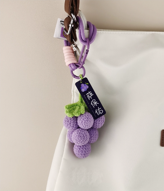 Cute purple grape-shaped knitted keychain, white bag with a tag featuring Chinese characters, product photography on a simple background, high-definition details.