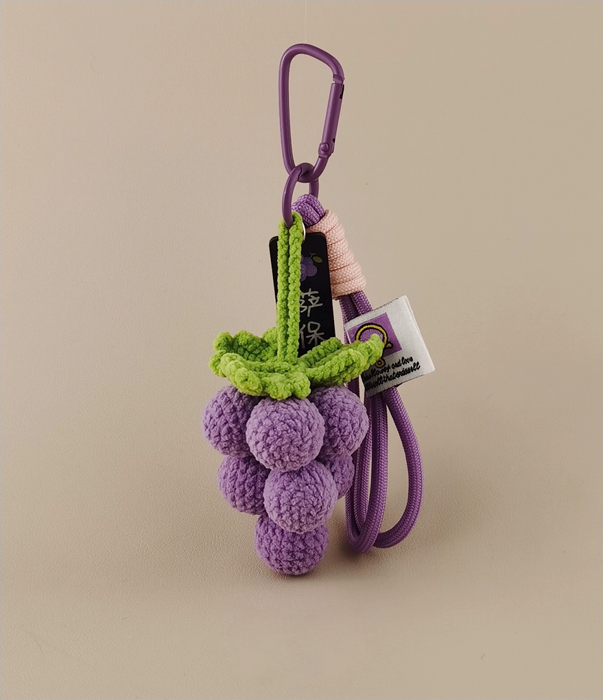 A purple grape-shaped keychain with a green vine, a white tag, and a lanyard, made of crocheted wool fabric and yarn material. The product photography features a simple, solid-colored light brown background, showcasing the purple grapes in a cute, high-definition style.