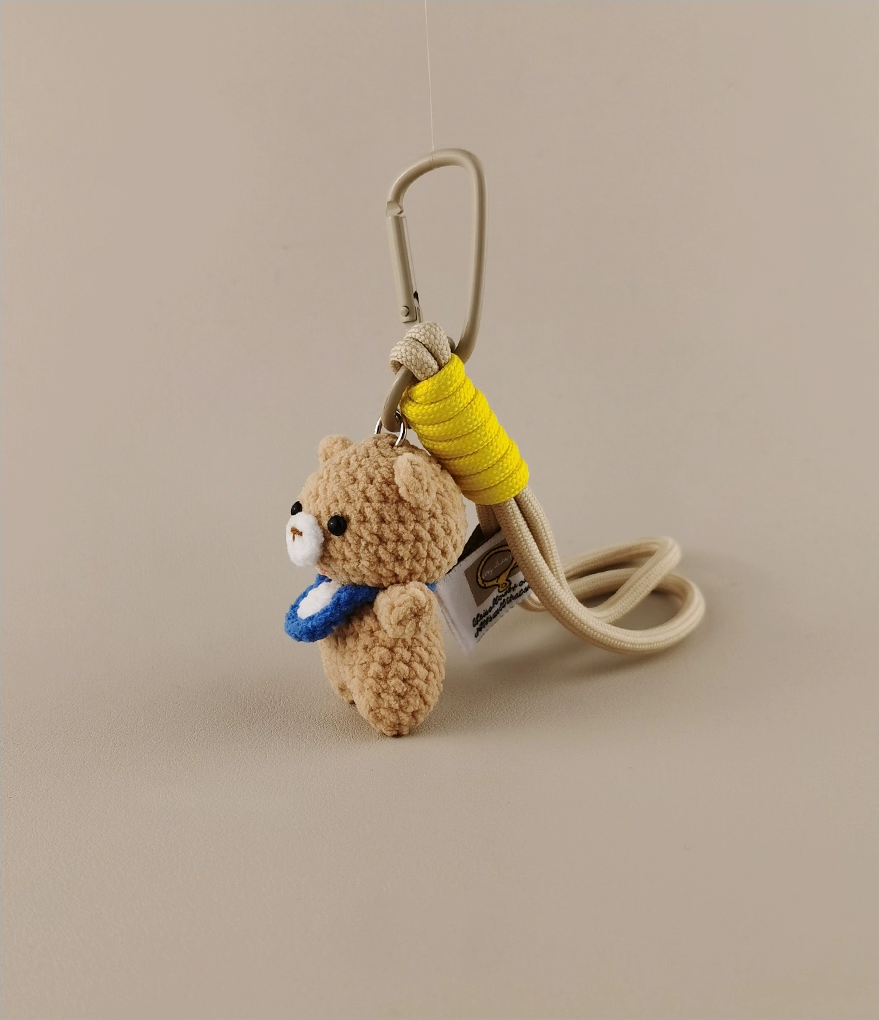 Simple and cute kawaii crochet bear with a blue neckerchief, holding a yellow jump rope as a keychain, against a light beige background, product photography