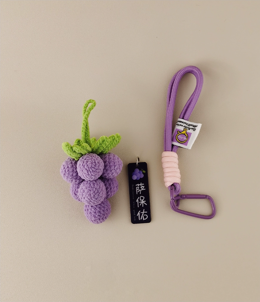 Cute, purple grape-shaped amigurumi keychain. Next to the black and white label is a long lanyard for carrying keys or small items. The pale lavender background sets off the vivid colors of the grapes. This design highlights an adorable fruit motif in vibrant hues, adding charm and playfulness to your bag. Perfect as a fun accessory!