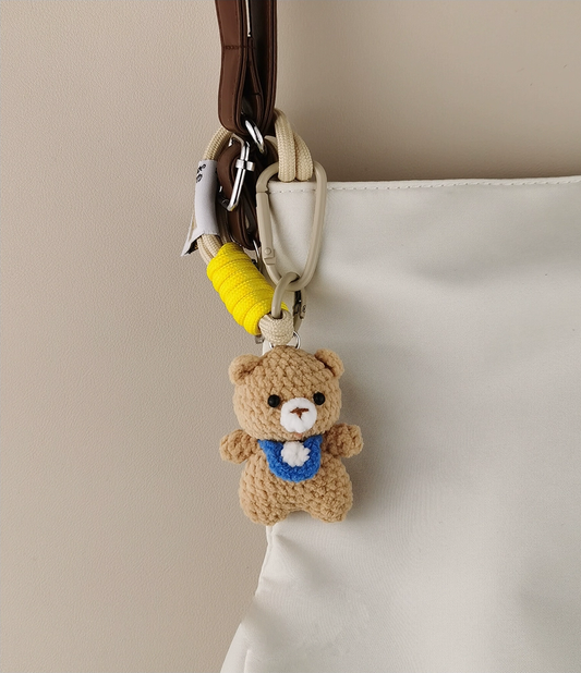 Simple, small crochet bear pendant in beige and blue, with a cartoon-cute shape. A white bag with a yellow hanging belt, against a solid color background, in the style of a cute plush toy.
