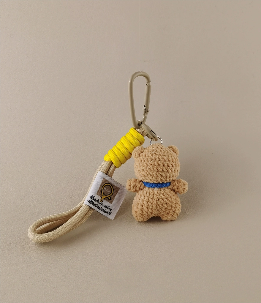 Simple keychain with a small crocheted beige teddy bear in a blue collar, a white tag, and a yellow carabiner on a light gray background. Minimalist style for handmade products for a children's store or home decor.