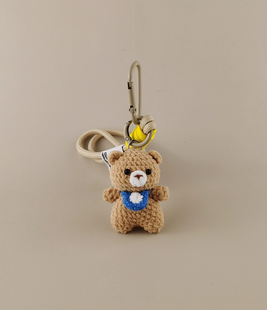 Simple cute bear keychain, with a blue collar and white belly patch in the style of Amigurumi. The background is beige. A yellow banana hangs on the hook of an anime bag or schoolbag, cartoon character design, high resolution, high detail, high quality.