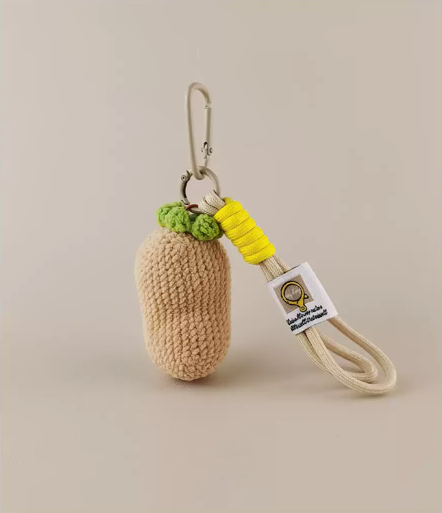 A knitted keychain in the shape of an eggplant, with a white tag and yellow laces with green tips attached to the bottom part. Product photography, studio lighting, and a beige background. -