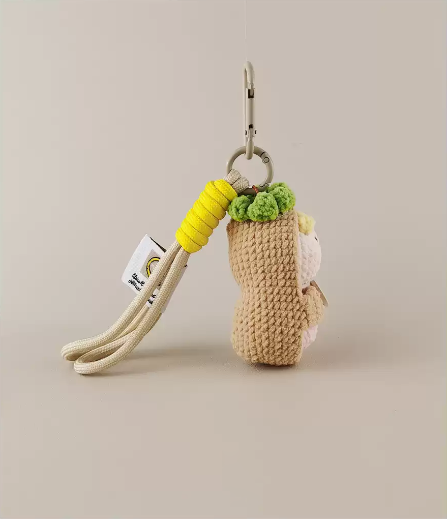 A keychain with an amigurumi cute little monkey, featuring a yellow and green color palette, made of yarn, against a white background, with a detailed character design, in the toycore and oshare kei styles. A long beige rope hangs down from the bottom.