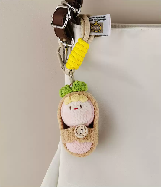Simple crochet keychain with a cute little doll inside, featuring beige and pink tones on a white background. A yellow ribbon adorns the top of the shoulder bag strap. The small baby toy is in an open-mouth pose, with a green leaves headband around its neck. The texture and fabric material are depicted in detail. A white cloth hangs from the bottom edge of the photo. High resolution.