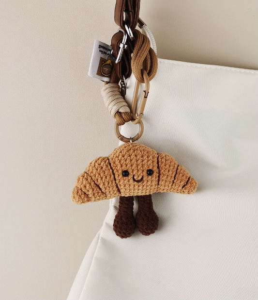 This is a crocheted keychain. It's designed to look like a cute croissant with a smiling face. The croissant has a light brown, textured appearance, black eyes, and a small curved smile. It also features two dark brown, cylindrical legs. Attached to it is a brown cord and a metal carabiner, making it suitable for hanging keys or decorating bags. 