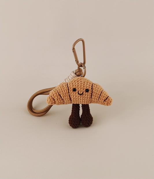 This is a crocheted keychain. It's designed to look like a cute croissant with a smiling face. The croissant has a light brown, textured appearance, black eyes, and a small curved smile. It also features two dark brown, cylindrical legs. Attached to it is a brown cord and a metal carabiner, making it suitable for hanging keys or decorating bags. 