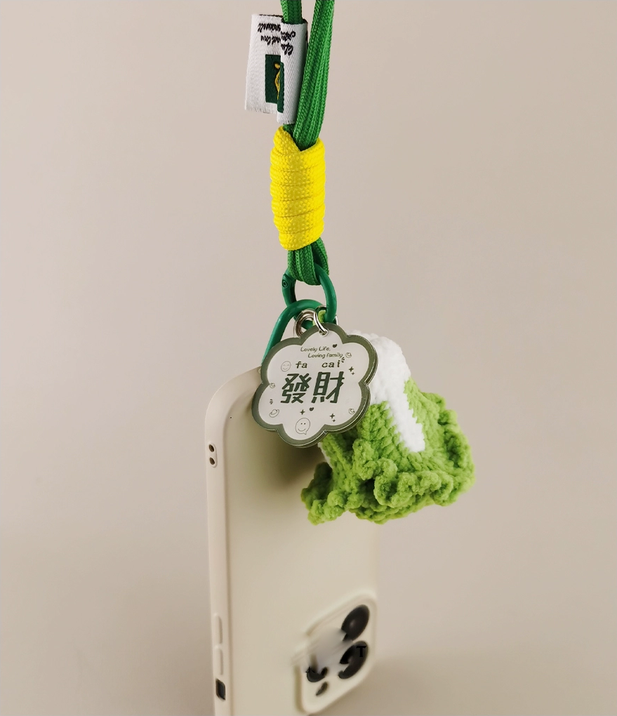 This is a crocheted keychain shaped like a Chinese cabbage. The cabbage has a white stem and green leaves, crafted with fine crochet work. It’s attached to a green carabiner and a green lanyard, with a small tag on the lanyard. The overall design is cute and vivid, making it a lovely accessory for keys or bags. 