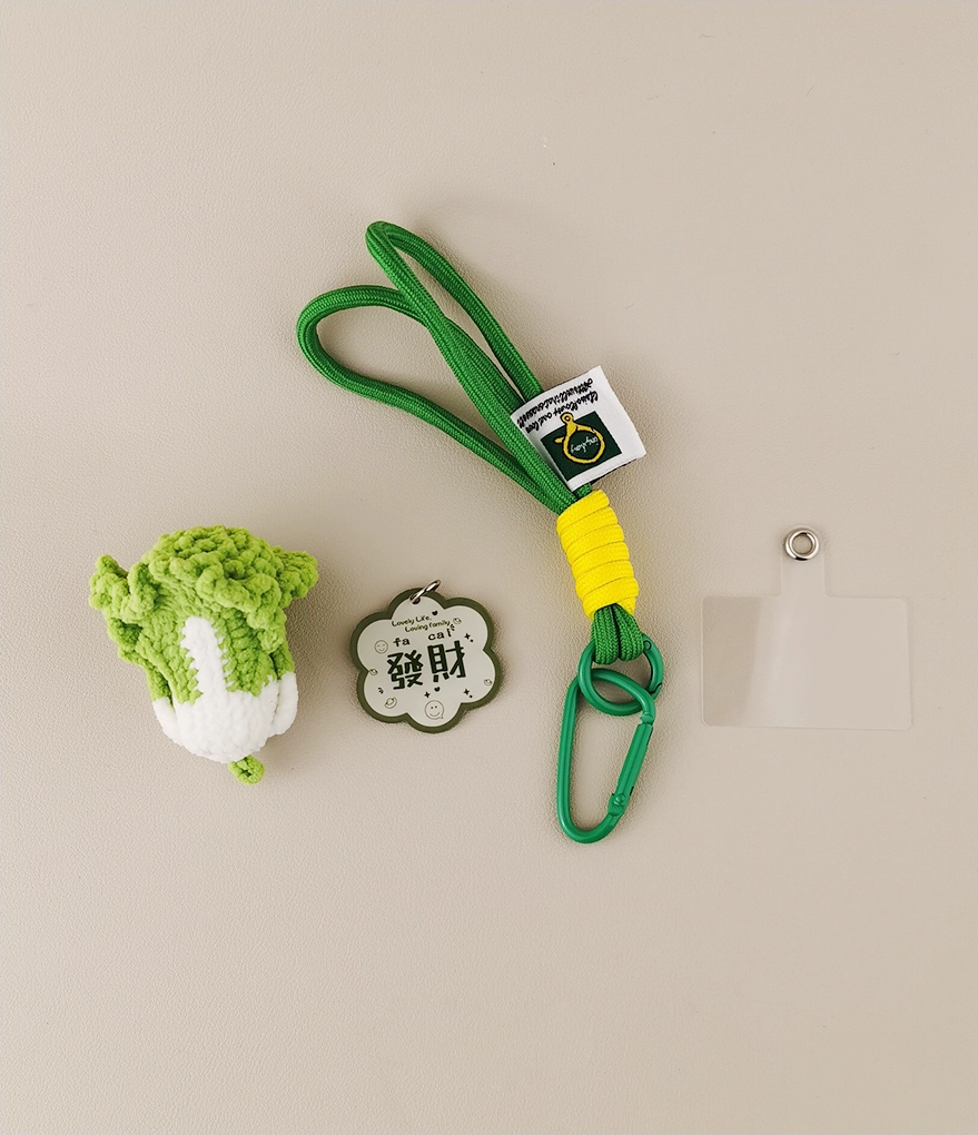 This is a crocheted keychain shaped like a Chinese cabbage. The cabbage has a white stem and green leaves, crafted with fine crochet work. It’s attached to a green carabiner and a green lanyard, with a small tag on the lanyard. The overall design is cute and vivid, making it a lovely accessory for keys or bags. 