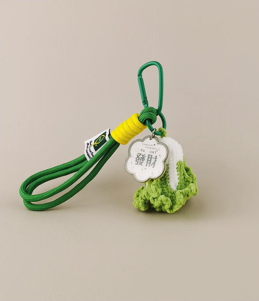 This is a crocheted keychain shaped like a Chinese cabbage. The cabbage has a white stem and green leaves, crafted with fine crochet work. It’s attached to a green carabiner and a green lanyard, with a small tag on the lanyard. The overall design is cute and vivid, making it a lovely accessory for keys or bags. 