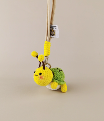 These are two crocheted keychains. One is a cute bee with a yellow and brown body, white wings, and yellow antennae. The other is a small green turtle - like figure with a green shell and yellow limbs, seemingly peeking out from a white "shell". They look very adorable and are nice little handicrafts. 