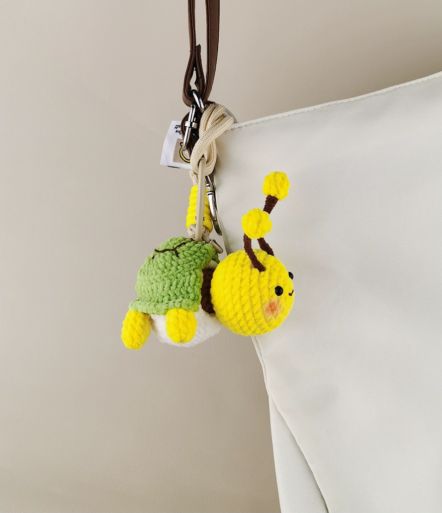 These are two crocheted keychains. One is a cute bee with a yellow and brown body, white wings, and yellow antennae. The other is a small green turtle - like figure with a green shell and yellow limbs, seemingly peeking out from a white "shell". They look very adorable and are nice little handicrafts. 