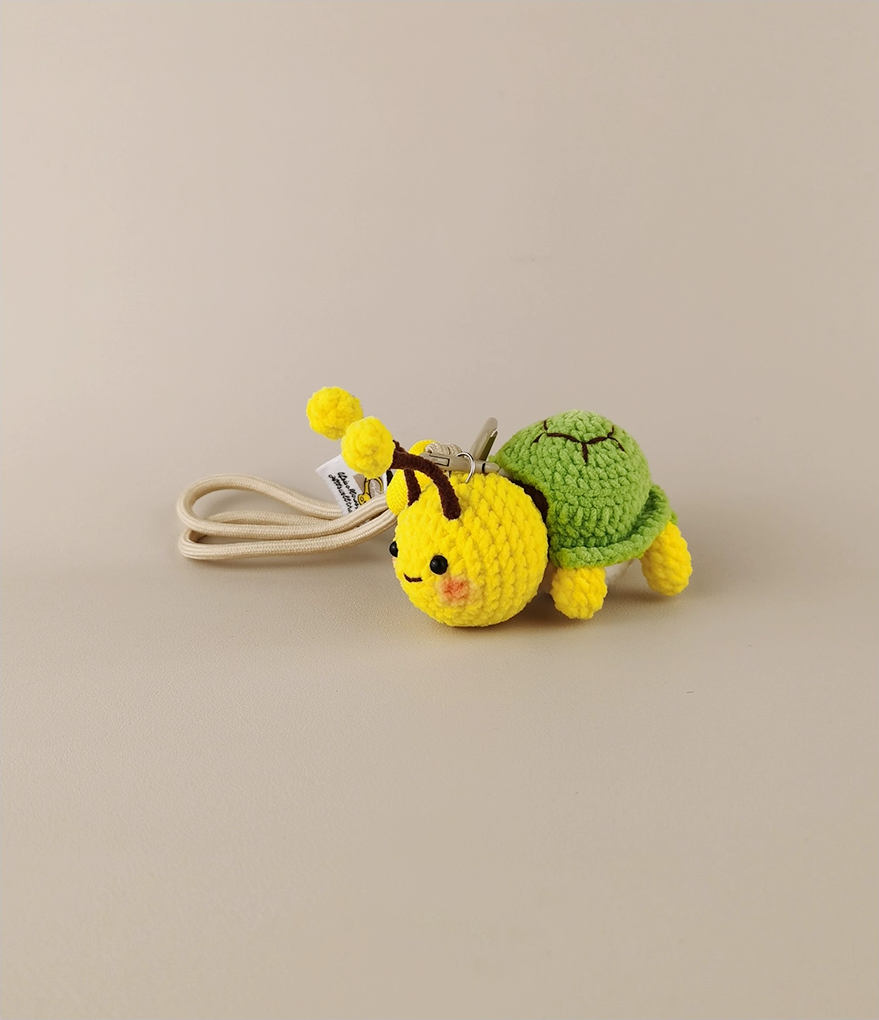 These are two crocheted keychains. One is a cute bee with a yellow and brown body, white wings, and yellow antennae. The other is a small green turtle - like figure with a green shell and yellow limbs, seemingly peeking out from a white "shell". They look very adorable and are nice little handicrafts. 