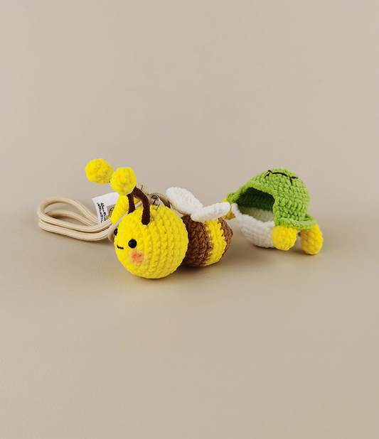 These are two crocheted keychains. One is a cute bee with a yellow and brown body, white wings, and yellow antennae. The other is a small green turtle - like figure with a green shell and yellow limbs, seemingly peeking out from a white "shell". They look very adorable and are nice little handicrafts. 