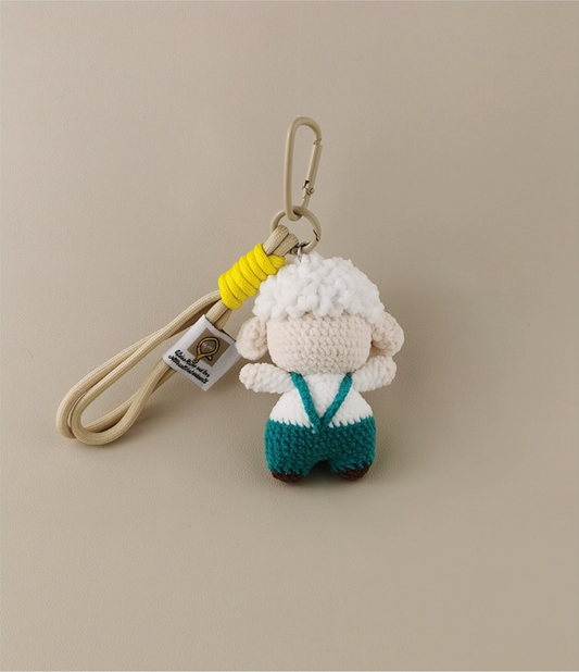 This is a crocheted keychain featuring a cute sheep character. The sheep has a fluffy white "wool" head, a light - colored face and body, and is dressed in teal overalls. It's attached to a beige lanyard with a yellow accent and a metal carabiner. There's also a small tag on the lanyard. The overall design is adorable and handmade - looking. 