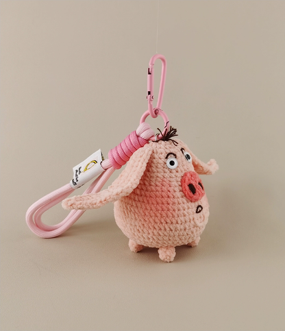 This is a crocheted keychain in the shape of a cute pink pig. The pig has large, expressive eyes, a round pink nose, and a small tuft of hair on its head. It's attached to a pink carabiner and a pink lanyard, with a small tag on the lanyard. The overall design is adorable and handmade - looking.