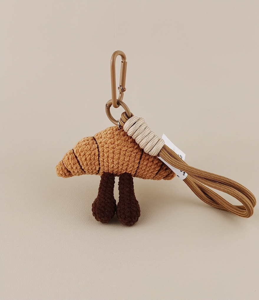 This is a crocheted keychain. It's designed to look like a cute croissant with a smiling face. The croissant has a light brown, textured appearance, black eyes, and a small curved smile. It also features two dark brown, cylindrical legs. Attached to it is a brown cord and a metal carabiner, making it suitable for hanging keys or decorating bags. 