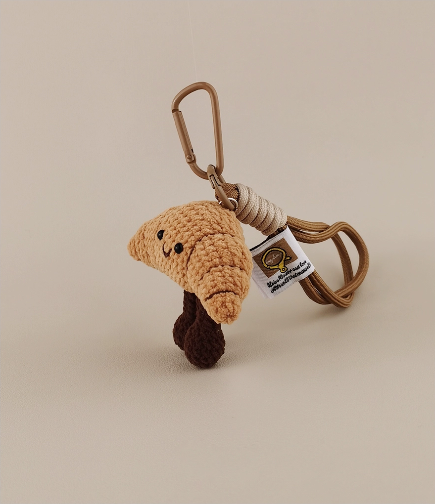 This is a crocheted keychain. It's designed to look like a cute croissant with a smiling face. The croissant has a light brown, textured appearance, black eyes, and a small curved smile. It also features two dark brown, cylindrical legs. Attached to it is a brown cord and a metal carabiner, making it suitable for hanging keys or decorating bags. 