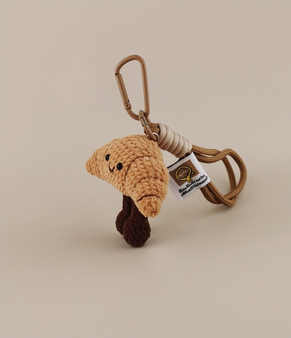 This is a crocheted keychain. It's designed to look like a cute croissant with a smiling face. The croissant has a light brown, textured appearance, black eyes, and a small curved smile. It also features two dark brown, cylindrical legs. Attached to it is a brown cord and a metal carabiner, making it suitable for hanging keys or decorating bags. 