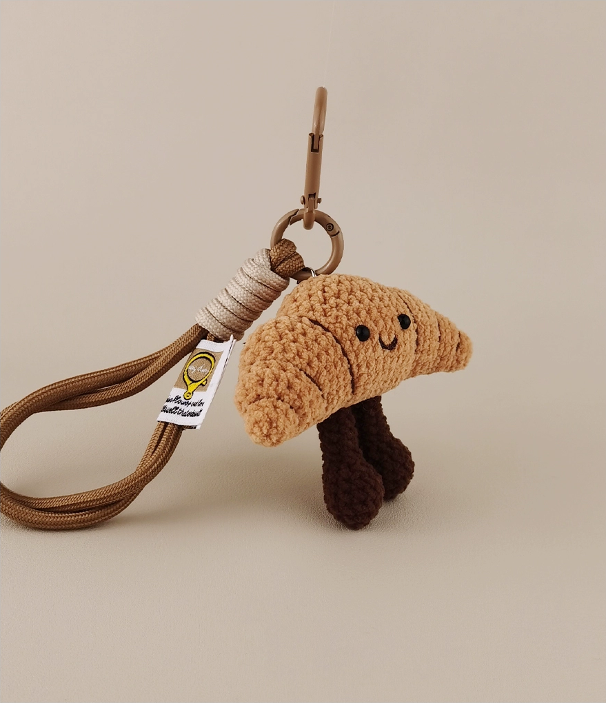 This is a crocheted keychain. It's designed to look like a cute croissant with a smiling face. The croissant has a light brown, textured appearance, black eyes, and a small curved smile. It also features two dark brown, cylindrical legs. Attached to it is a brown cord and a metal carabiner, making it suitable for hanging keys or decorating bags. 