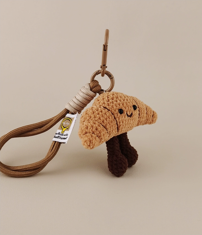 This is a crocheted keychain. It's designed to look like a cute croissant with a smiling face. The croissant has a light brown, textured appearance, black eyes, and a small curved smile. It also features two dark brown, cylindrical legs. Attached to it is a brown cord and a metal carabiner, making it suitable for hanging keys or decorating bags. 