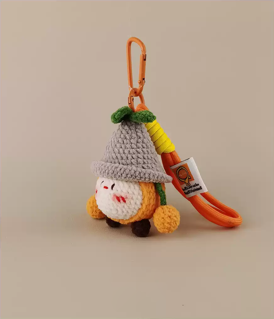 A reliance on amigurumi keychain, a cute little orange fruit character with a hat and green leafy hair holding a yellow arrow that says "Sewing", a cute crochet toy with a light grey background, a white label tag, and an orange felt for hanging. 