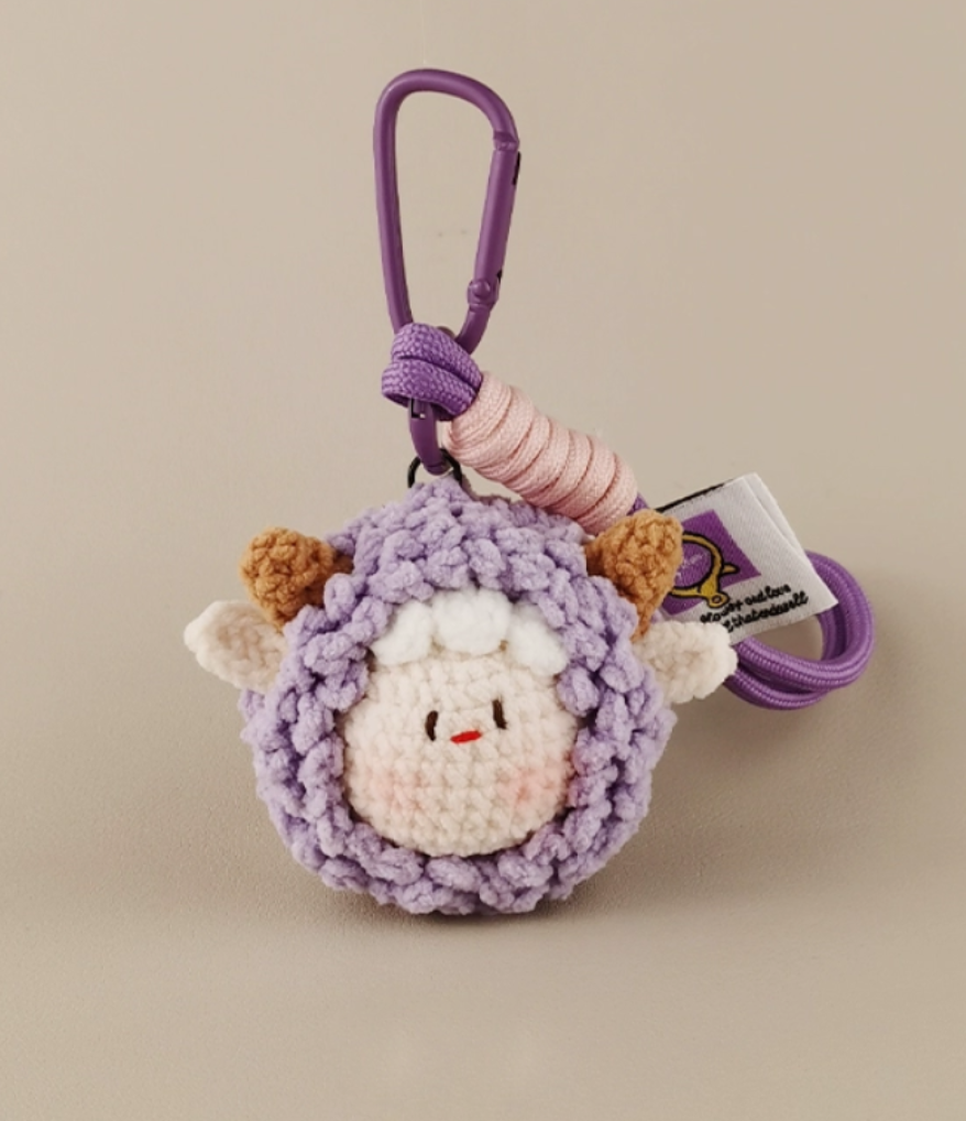 A purple crocheted keychain with an animal head, a white sheep face in the middle of it, with pink and brown hair on top, and a "St. Chiaro" tag attached to its tail. The background is light gray, with the character's posture facing right at eye level. It has a simple style, with soft lighting and a clear focus. 