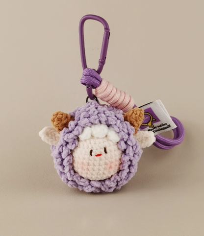 A purple crocheted keychain with an animal head, a white sheep face in the middle of it, with pink and brown hair on top, and a "St. Chiaro" tag attached to its tail. The background is light gray, with the character's posture facing right at eye level. It has a simple style, with soft lighting and a clear focus. 