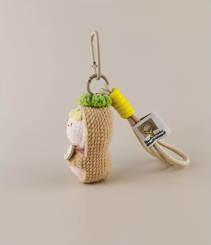 A small amigurumi keychain of an anthropomorphic potato with long hair, wearing a green leaf hat, a yellow string around the neck, and a white tag. The bottom is a brown, cream-colored, cylindrical shape made from crochet, with a simple design and a cute character. The background is detailed, and the product photography is well-executed.