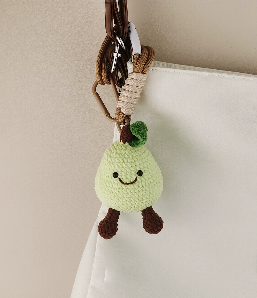 A cute, green pear-shaped character, a crocheted keychain hanging on the white bag with a brown handle. 