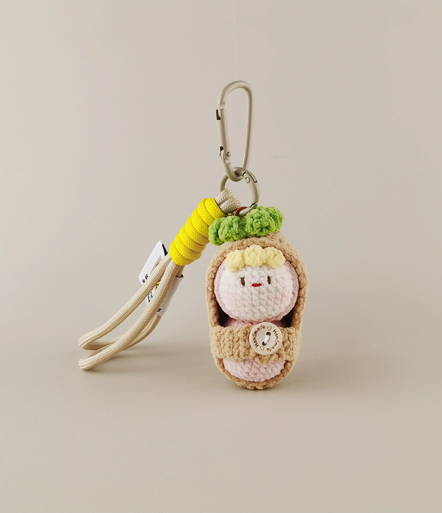 A keychain with an amigurumi, a cute baby in swaddling , featuring a light pink and beige color palette, with accents of yellow, white, green, and brown, made of yarn. On the top, there is a clip for carrying it around your bag or backpack, set against a white background, in the style of product photography. 