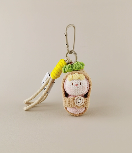 A keychain with an amigurumi, a cute baby in swaddling , featuring a light pink and beige color palette, with accents of yellow, white, green, and brown, made of yarn. On the top, there is a clip for carrying it around your bag or backpack, set against a white background, in the style of product photography. 