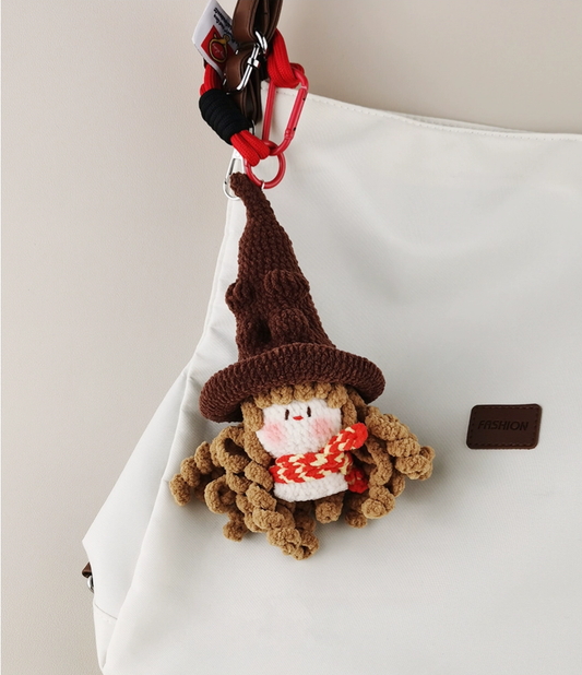  A cute little knitted witch with hat and scarf, made of brown yarn on the bag charm, is hanging from an airbrushed white totebag in full view. The totelust has "FASHION_awards" written on it. A red carlengine clip attached to its cap plastic headband was also visible.
