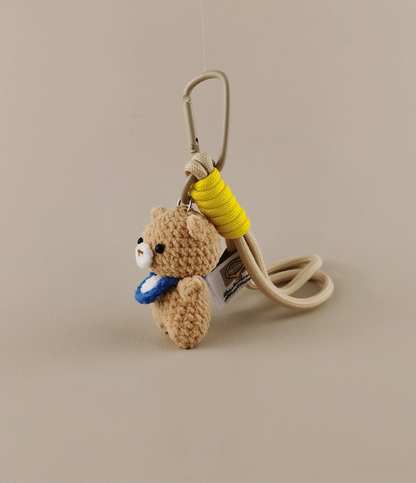 Simple and cute kawaii crochet bear with a blue neckerchief, holding a yellow jump rope as a keychain, against a light beige background, product photography