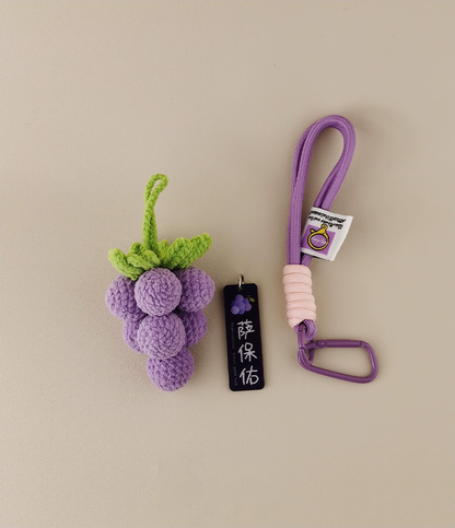 Cute, purple grape-shaped amigurumi keychain. Next to the black and white label is a long lanyard for carrying keys or small items. The pale lavender background sets off the vivid colors of the grapes. This design highlights an adorable fruit motif in vibrant hues, adding charm and playfulness to your bag. Perfect as a fun accessory! 