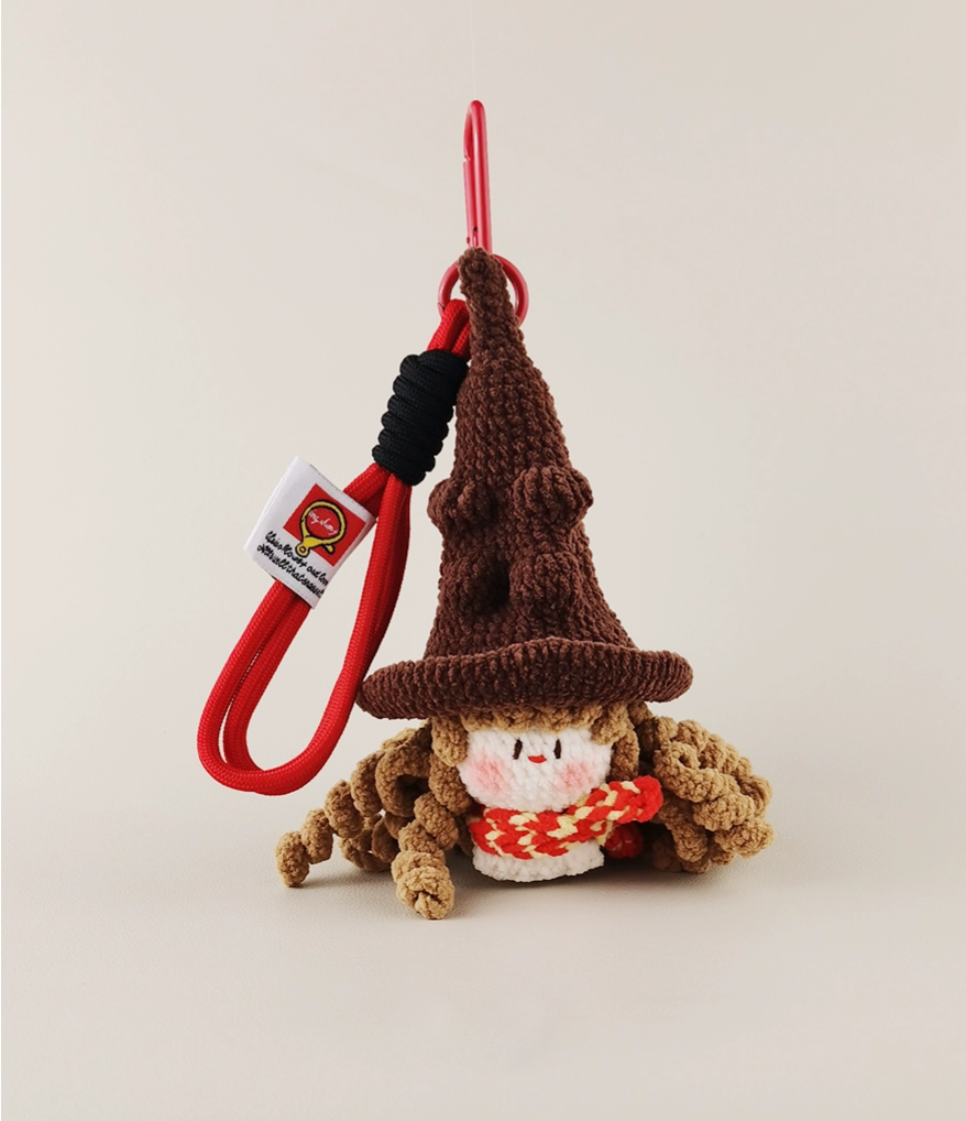 A very cute little brown wizard with a hat and red scarf is made of yarn, knitted in the shape of an amulet charm on a long red lanyard with a white tag. The background color should be a pure cream. A red line connected to its feet has been woven through by hand from the top left corner to the bottom right corner of the photo.
