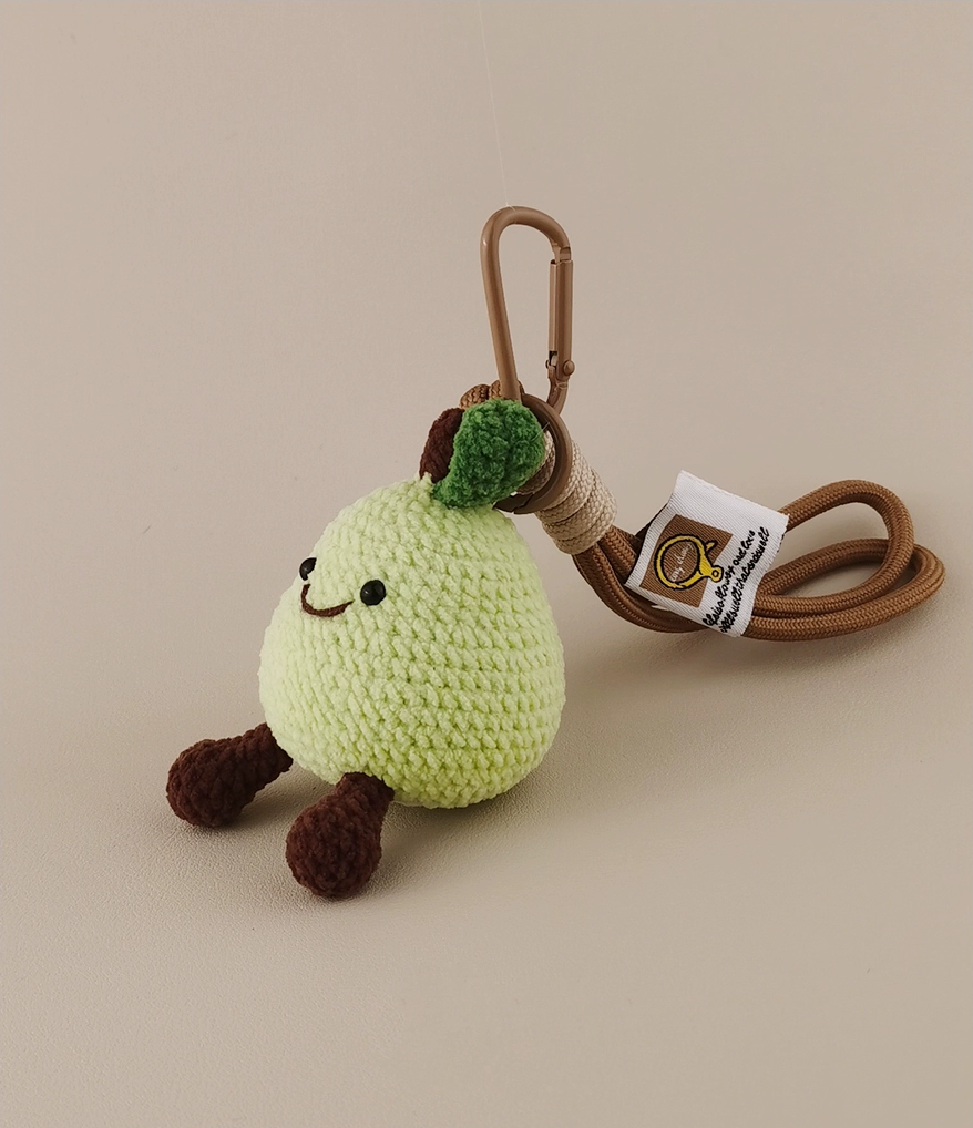 A cute little green pear with brown arms and legs, hand-crocheted in the style of amigurumi, has its head on the cartoon keychain attached to its body. There is also a tag label hanging from the end of that chain which contains some information about the design being used in these items. The background color should be light beige. 