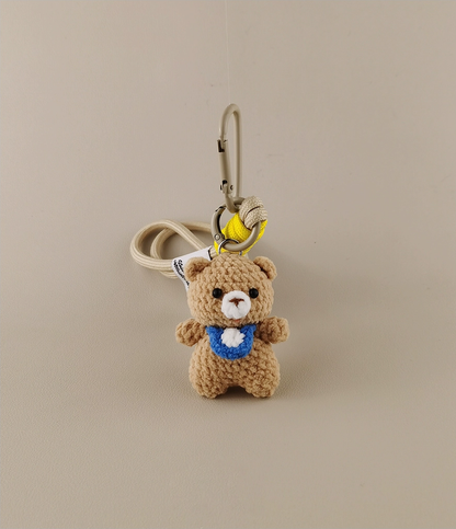 Simple cute bear keychain, with a blue collar and white belly patch in the style of Amigurumi. The background is beige. A yellow banana hangs on the hook of an anime bag or schoolbag, cartoon character design, high resolution, high detail, high quality.